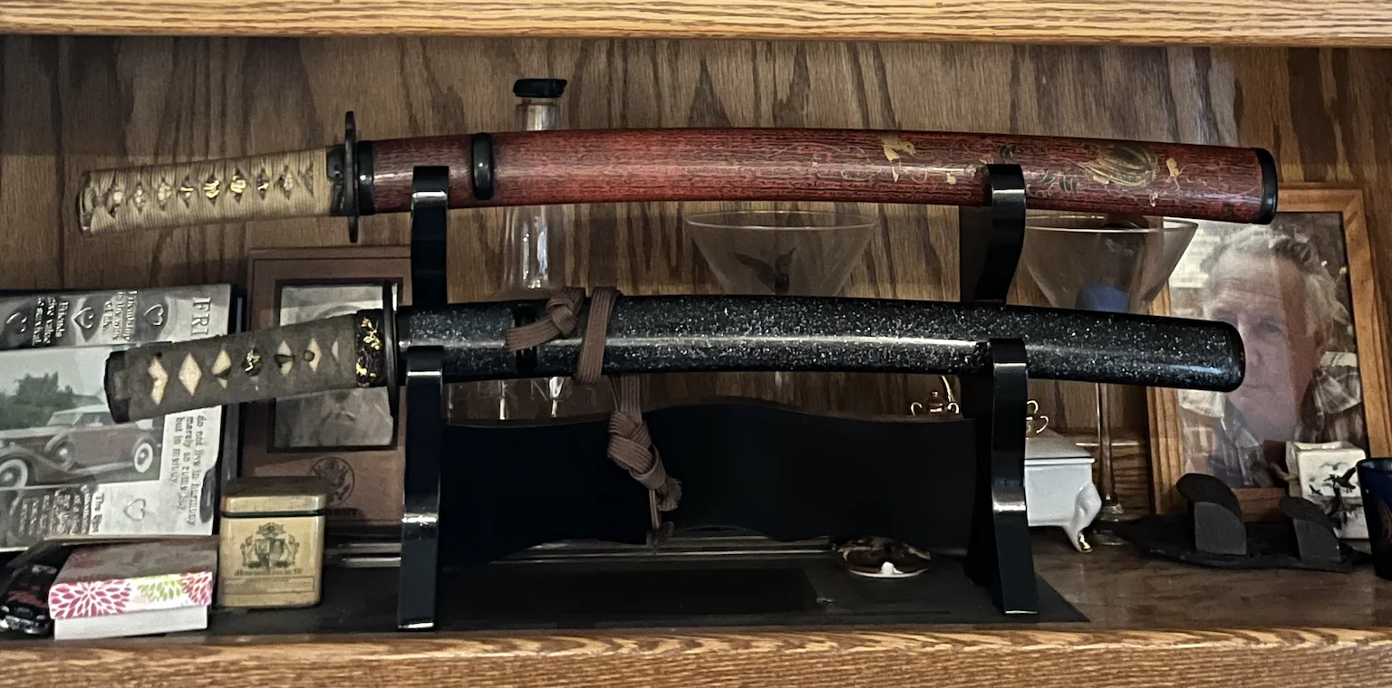Two sheathed Japanese swords displayed on a black lacquer stand, one in red lacquer koshirae and one in dark speckled koshirae.